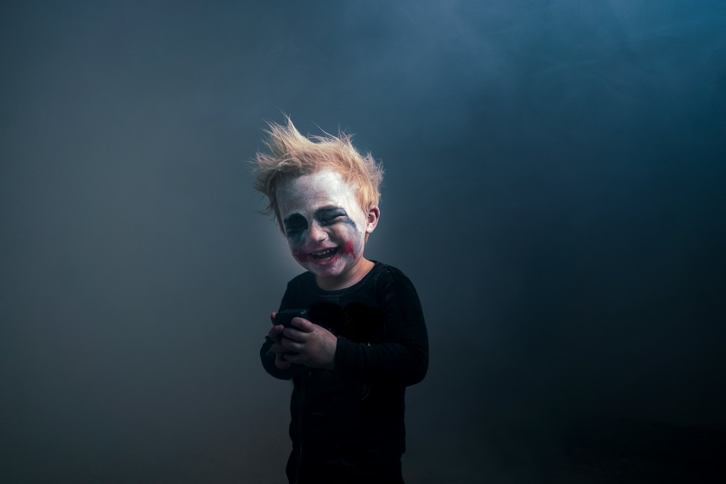 Child with Joker face paint smiling holding a dark object.
