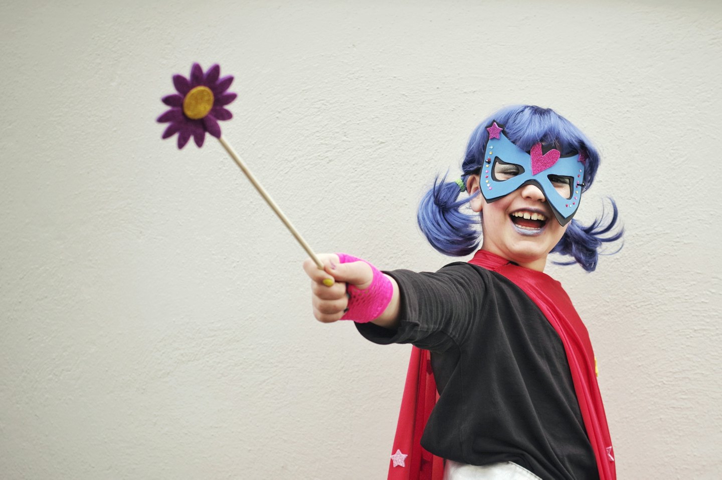 Child with blue hair wearing a superhero mask and cape holds a wand.